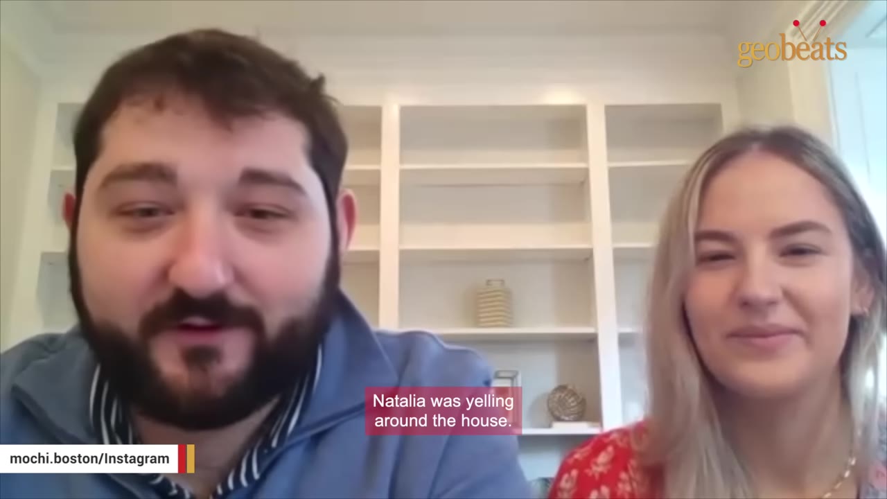 A man with a beard and a woman with blonde hair are sitting side-by-side in front of a white bookshelf, smiling during an interview.