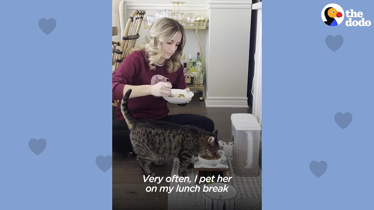 A woman sits on the floor eating from a white bowl while a tabby cat eats from its own bowl next to her.