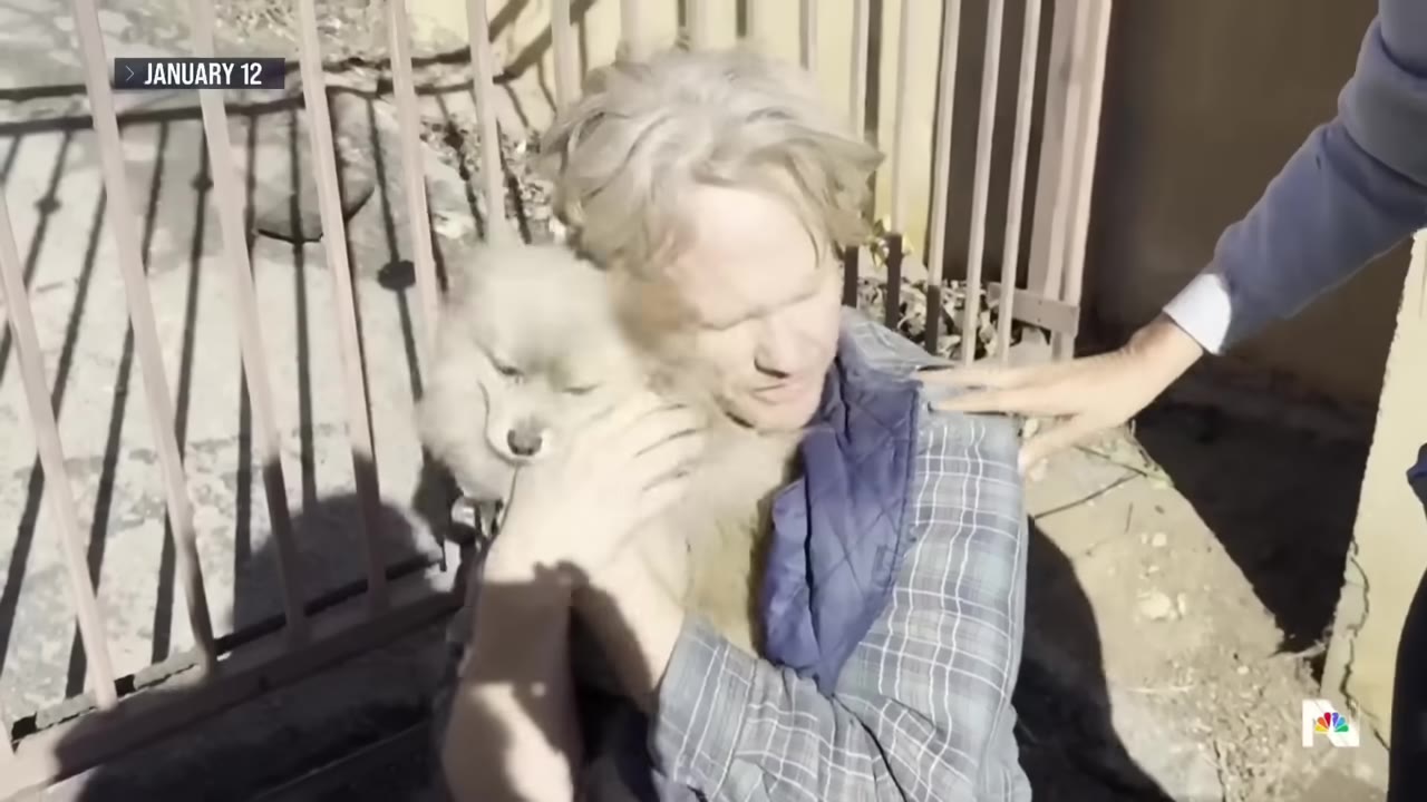 A man with blonde hair holds a small, fluffy light-colored dog close to his chest with an emotional expression, while another person places a comforting hand on his shoulder.