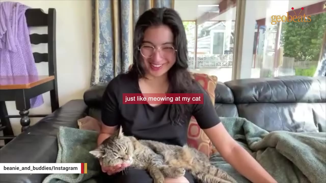 A smiling young woman with glasses sits on a couch, cradling a tabby cat in her lap.