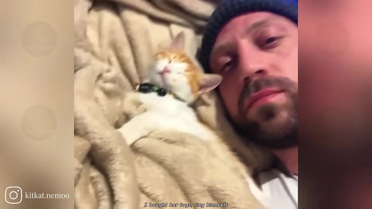 A man wearing a dark beanie lies down next to a small orange and white kitten sleeping comfortably on a soft beige blanket.