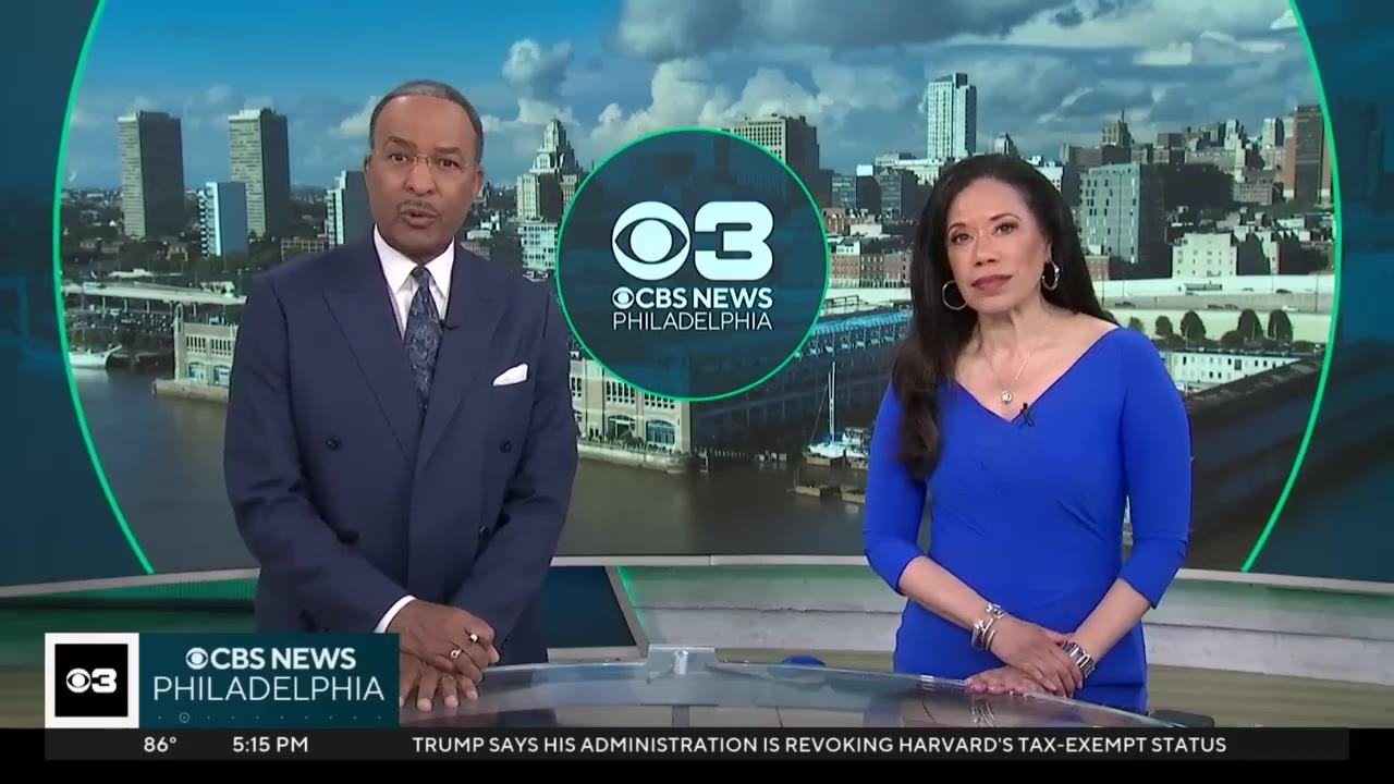 Two CBS News Philadelphia anchors, a man in a dark suit and a woman in a blue dress, stand behind a news desk looking directly at the camera.
