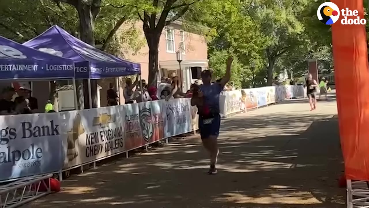 A man running down a marathon course lined with banners and tents, waving his hand as he approaches the finish area.