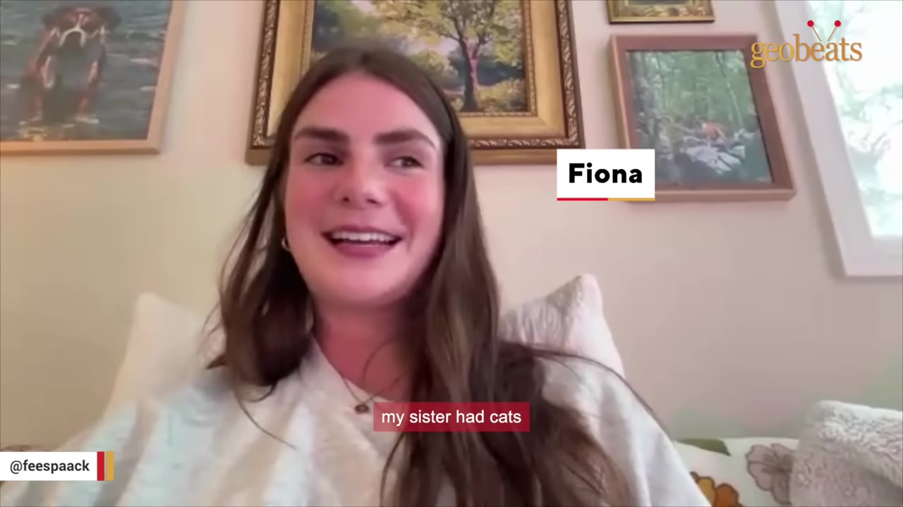 A young woman named Fiona with long brown hair smiles while speaking during an interview, with framed pictures on the wall behind her.