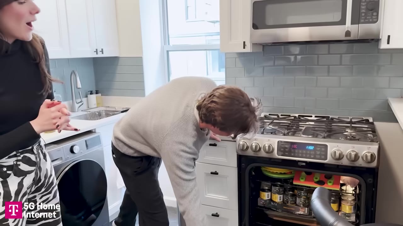A man in a grey sweater bends down to look inside an open oven filled with cat food cans and colorful pet toys, while a woman stands beside him in a kitchen.