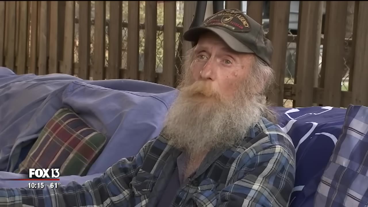 An older man with a long grey beard, wearing a plaid shirt and a cap, sitting outdoors.