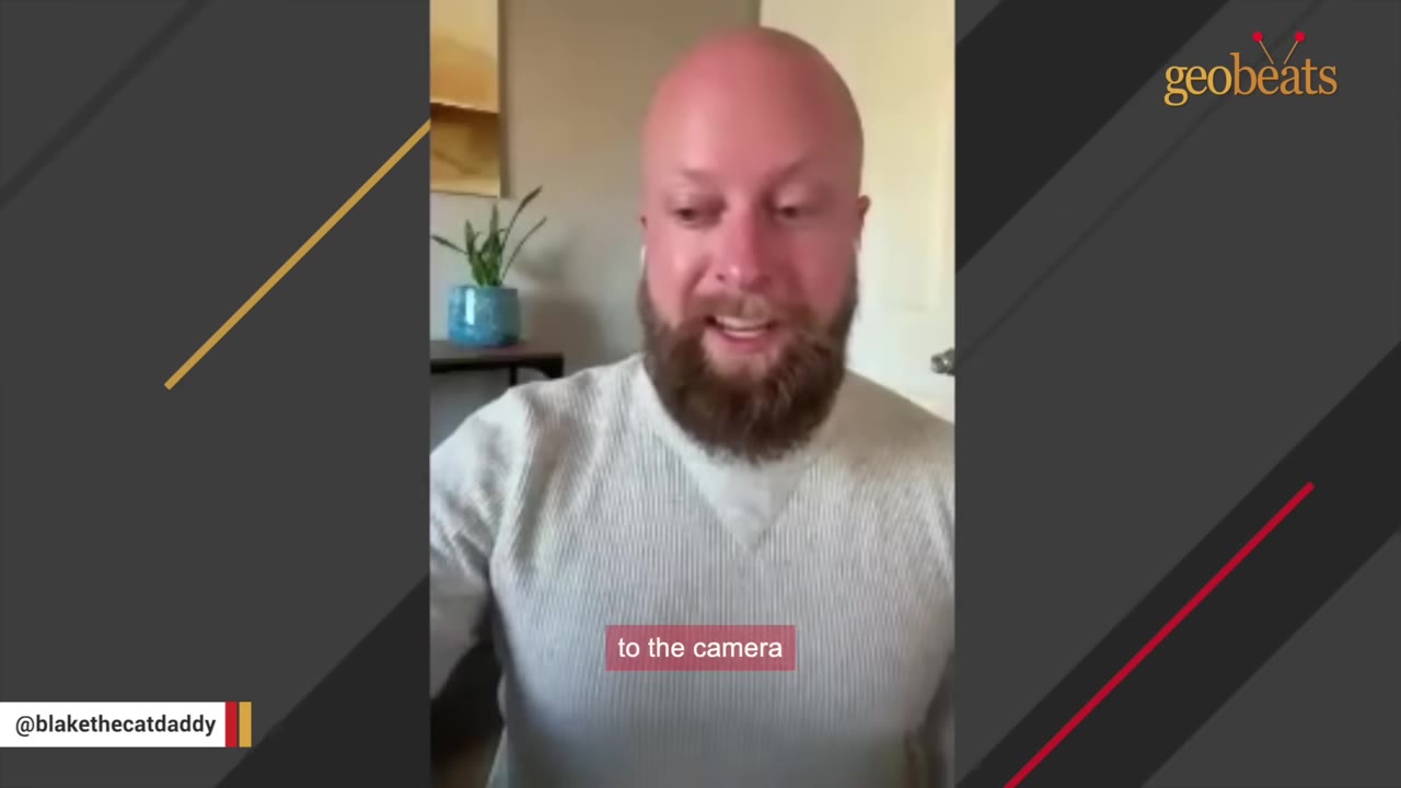 A bald man with a beard wearing a grey sweater speaks directly to the camera.