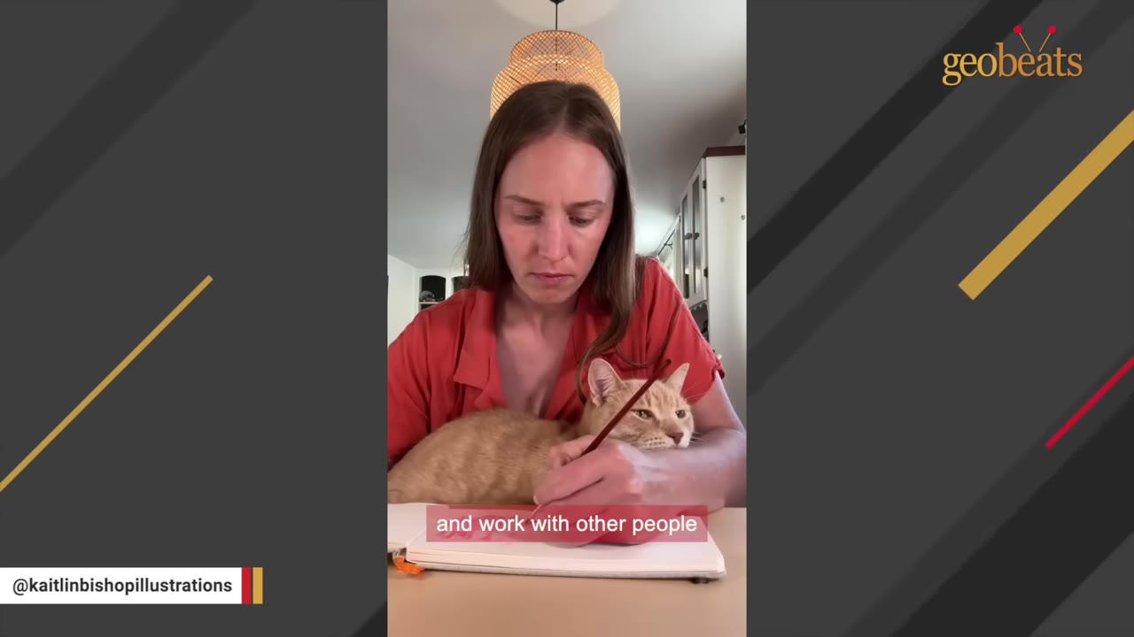A woman in a red shirt draws in a sketchbook with a paintbrush while an orange cat rests in her arms on the desk.