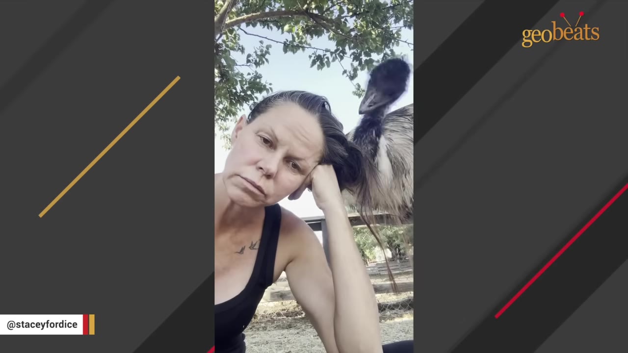 A large emu stands tall behind a woman who is resting her head on her hand.