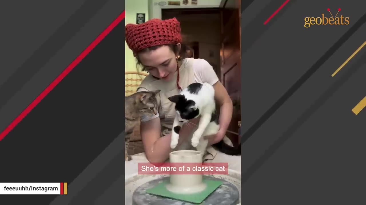 A woman wearing a red crocheted hat holds a black and white cat over a pottery wheel while a tabby cat sits nearby watching.
