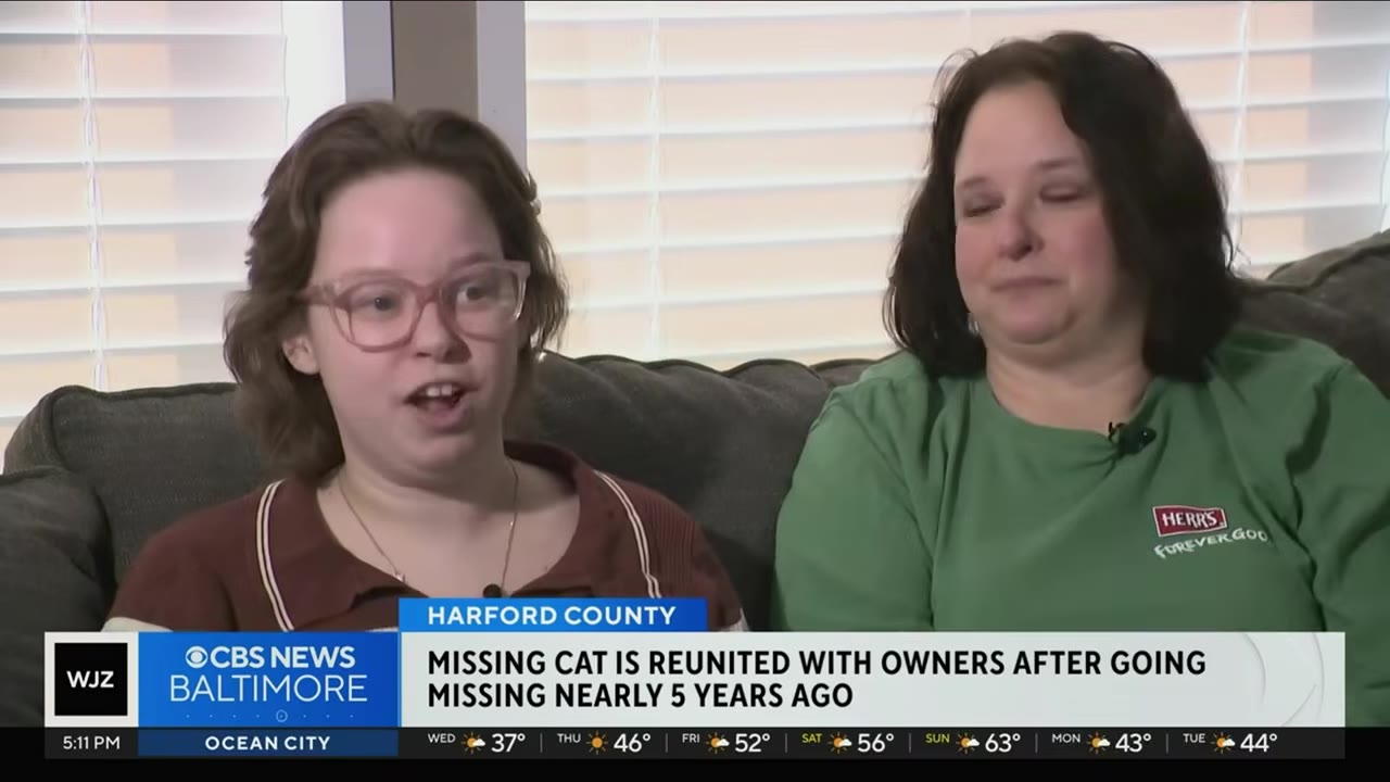 Two women sit on a couch during a news interview, one wearing glasses and speaking, while the other looks on with an emotional, tearful expression.