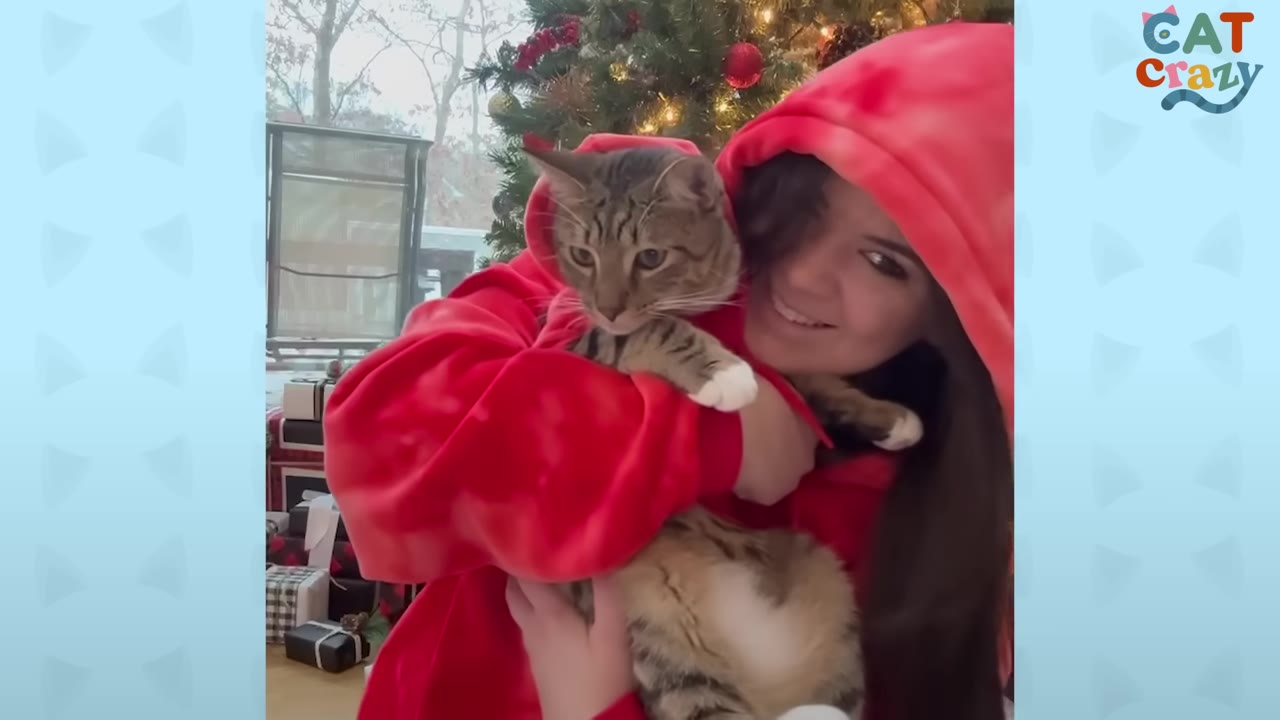 A smiling woman in a red hooded sweatshirt hugs a tabby cat who is also wearing a matching red hoodie, in front of a decorated Christmas tree.