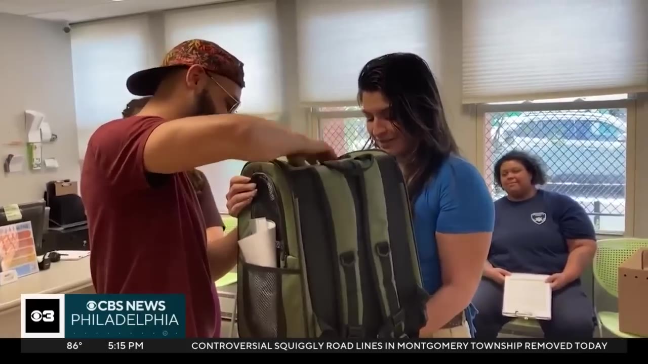 A man in a maroon shirt and a backward baseball cap and a woman in a blue shirt stand together in an office setting, looking at a green pet carrier backpack.