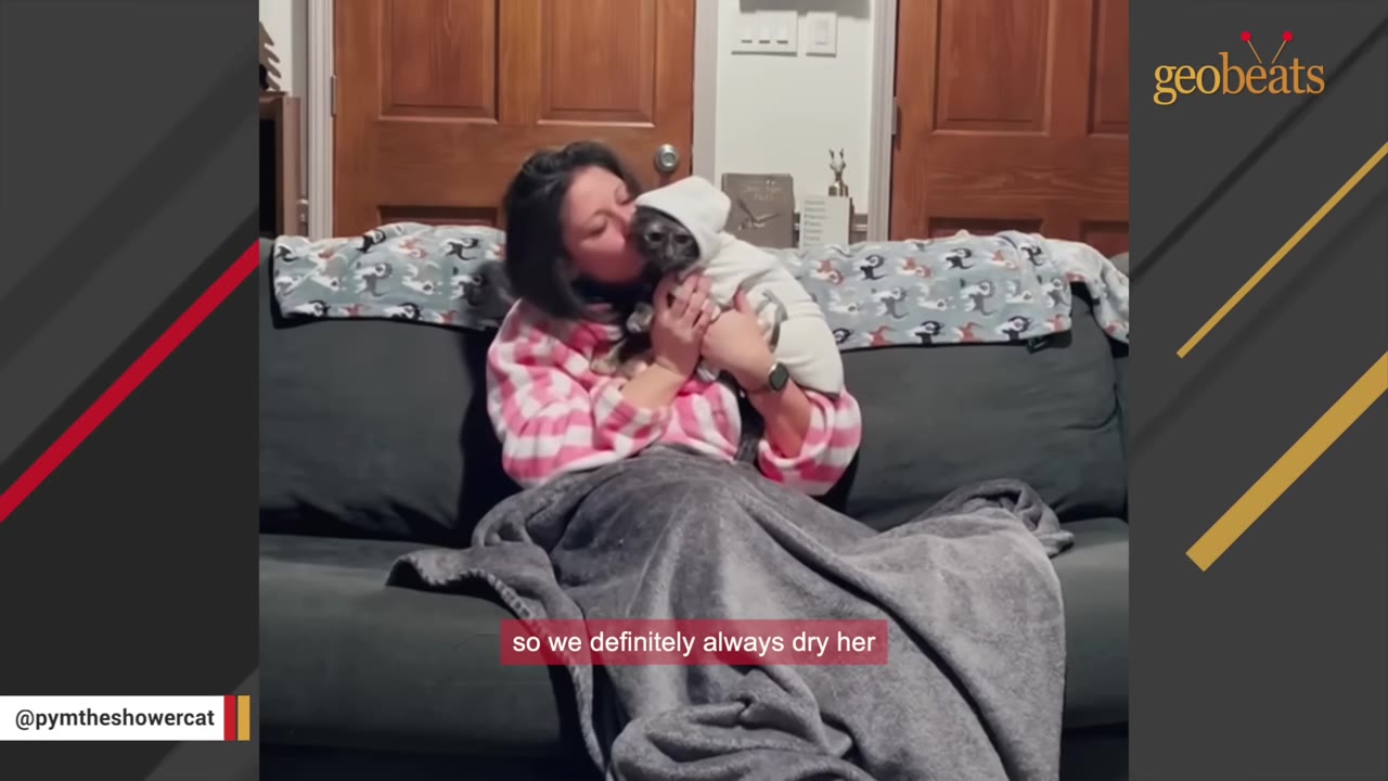 A woman in a pink and white striped robe sits on a couch holding a small dark cat wrapped in a white hooded towel.