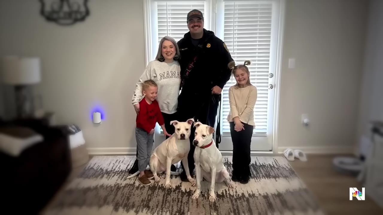 A family of four, including a mother, a father in a uniform, and two young children, pose together indoors with two white dogs sitting in front of them.