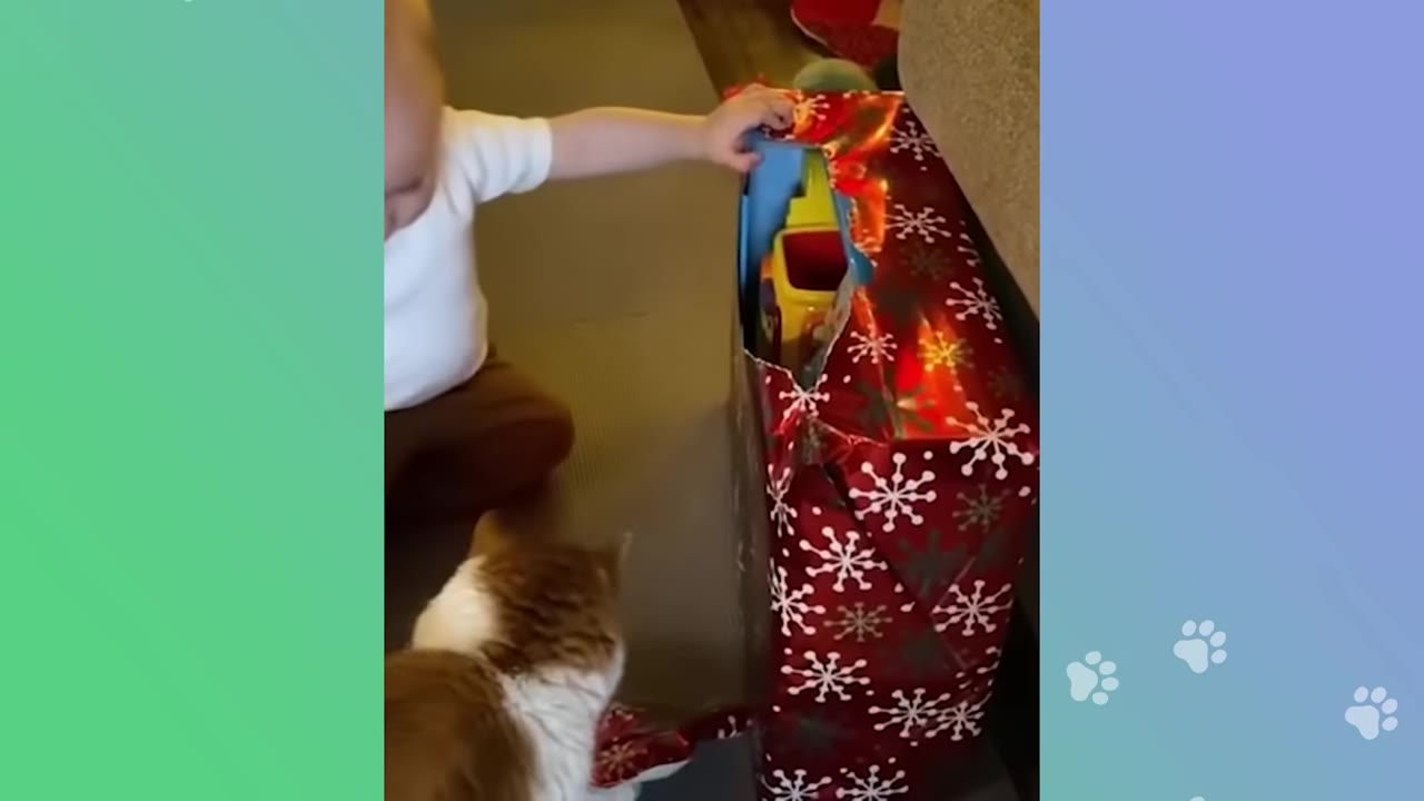 A toddler in a white shirt sits on the floor next to an orange and white cat, reaching towards a large gift wrapped in red paper with snowflake patterns.