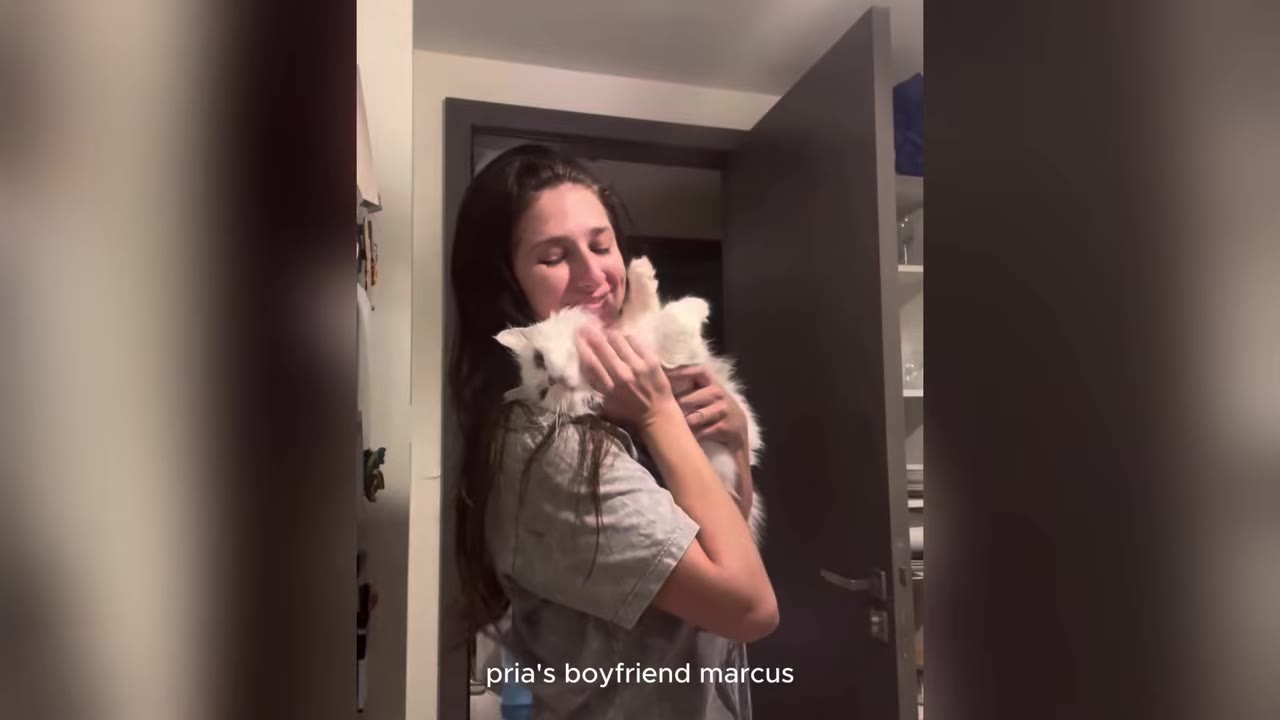 A woman with long dark hair smiles as she holds a fluffy white kitten close to her chest in a doorway.