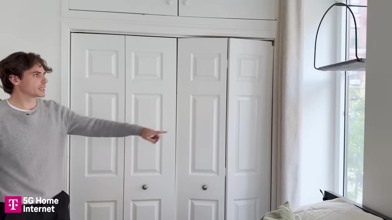 A man in a grey sweater points towards white closet doors and overhead storage cabinets in a bedroom.