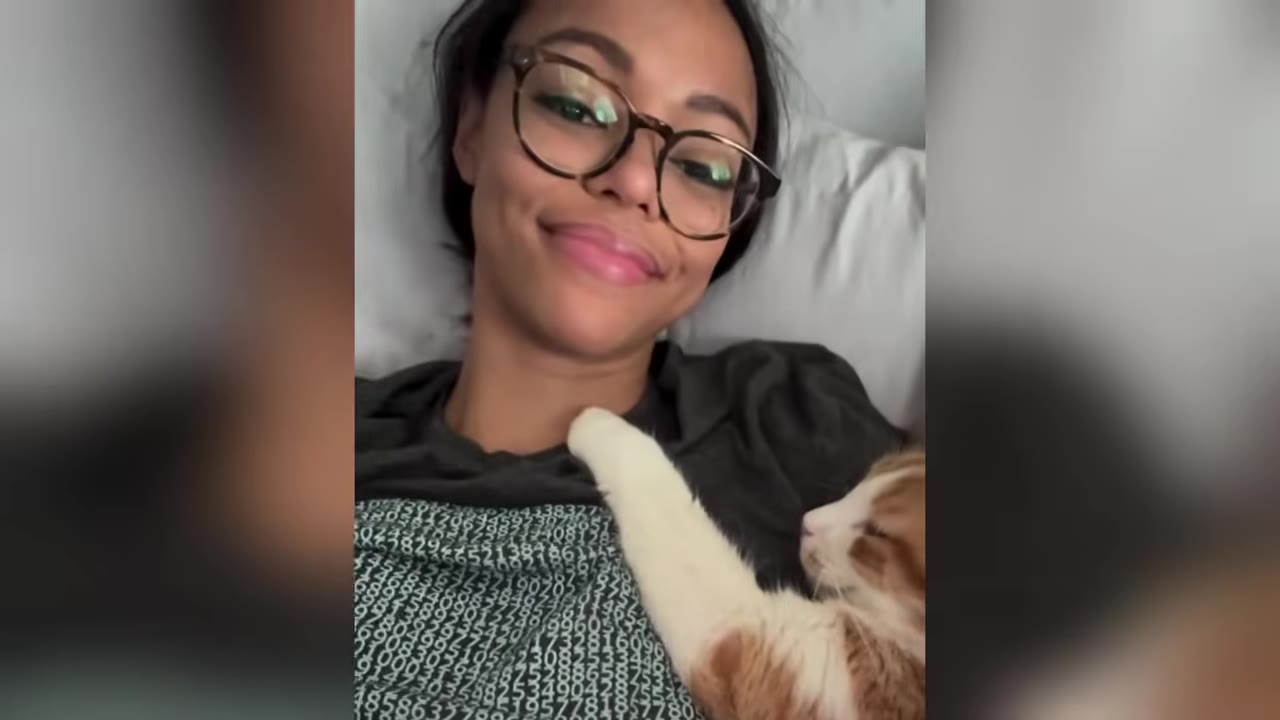 A smiling woman wearing glasses lies down with an orange and white cat resting its paw on her chest.