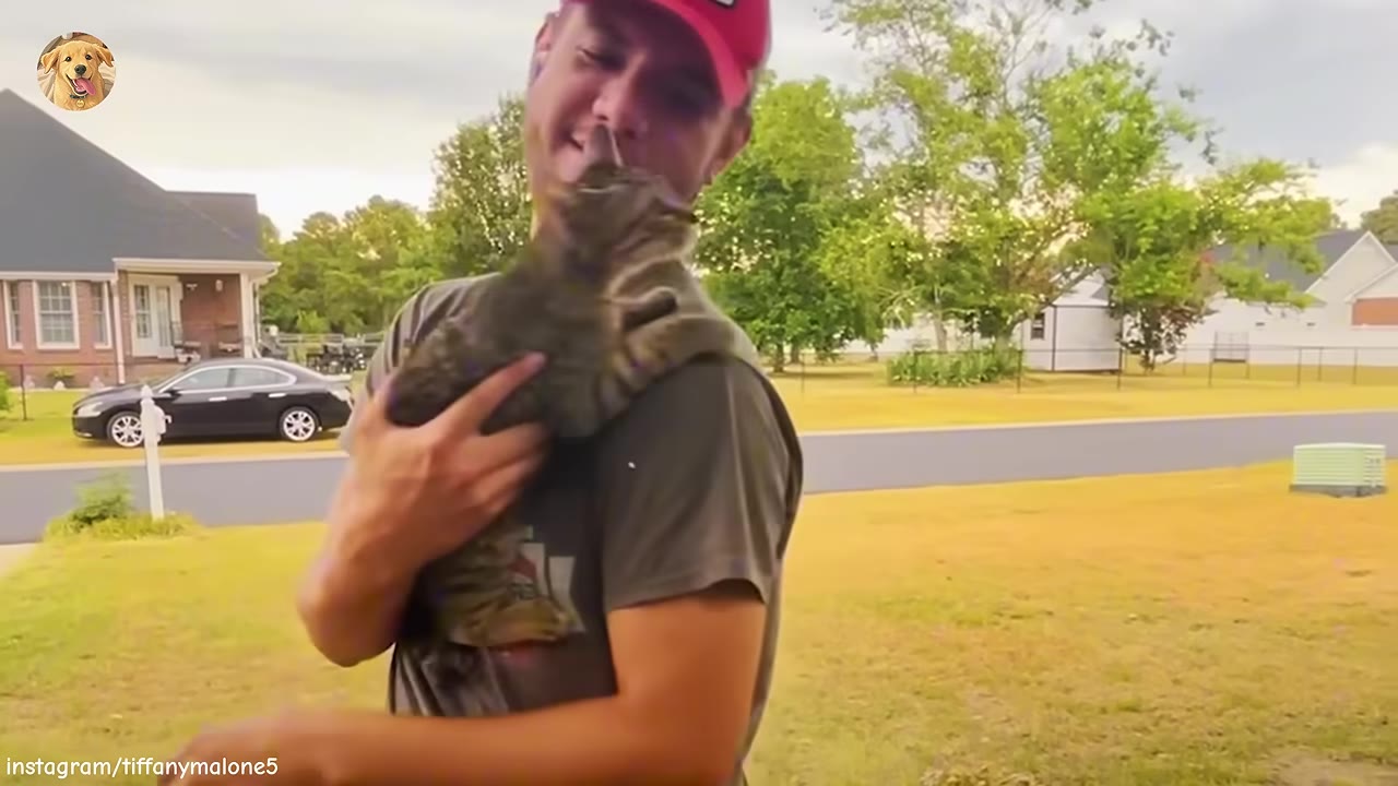 A smiling man in a red cap holding a tabby cat close to his shoulder outdoors.