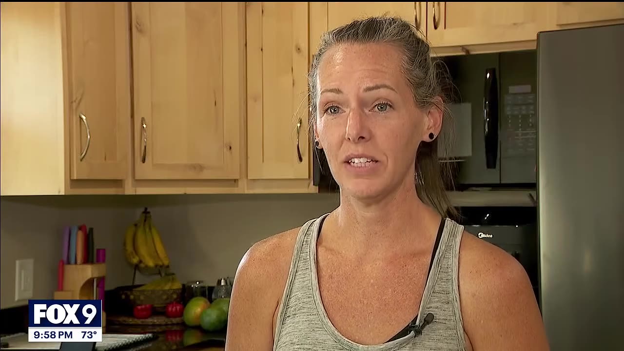A woman with grey-streaked hair tied back, wearing a grey tank top, speaking in a kitchen with wooden cabinets.