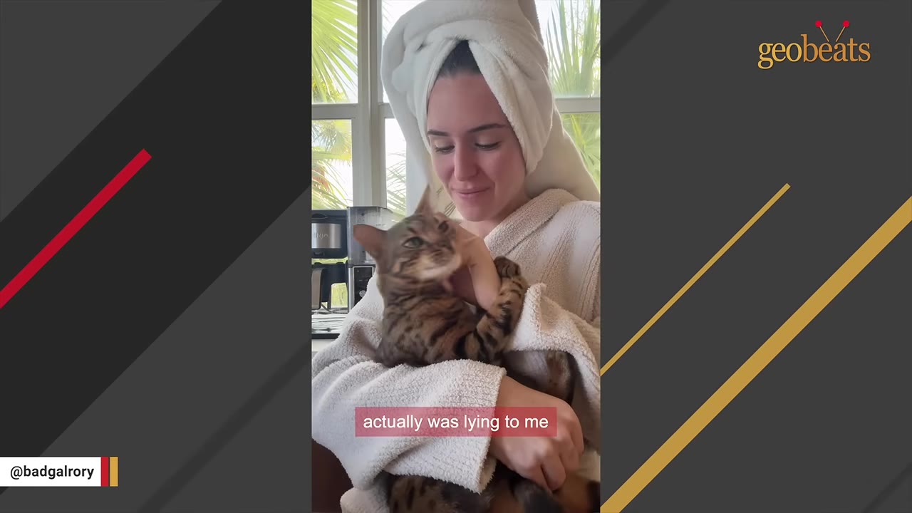 A woman with a white towel wrapped around her head holds a brown tabby cat in her arms while looking down at it.