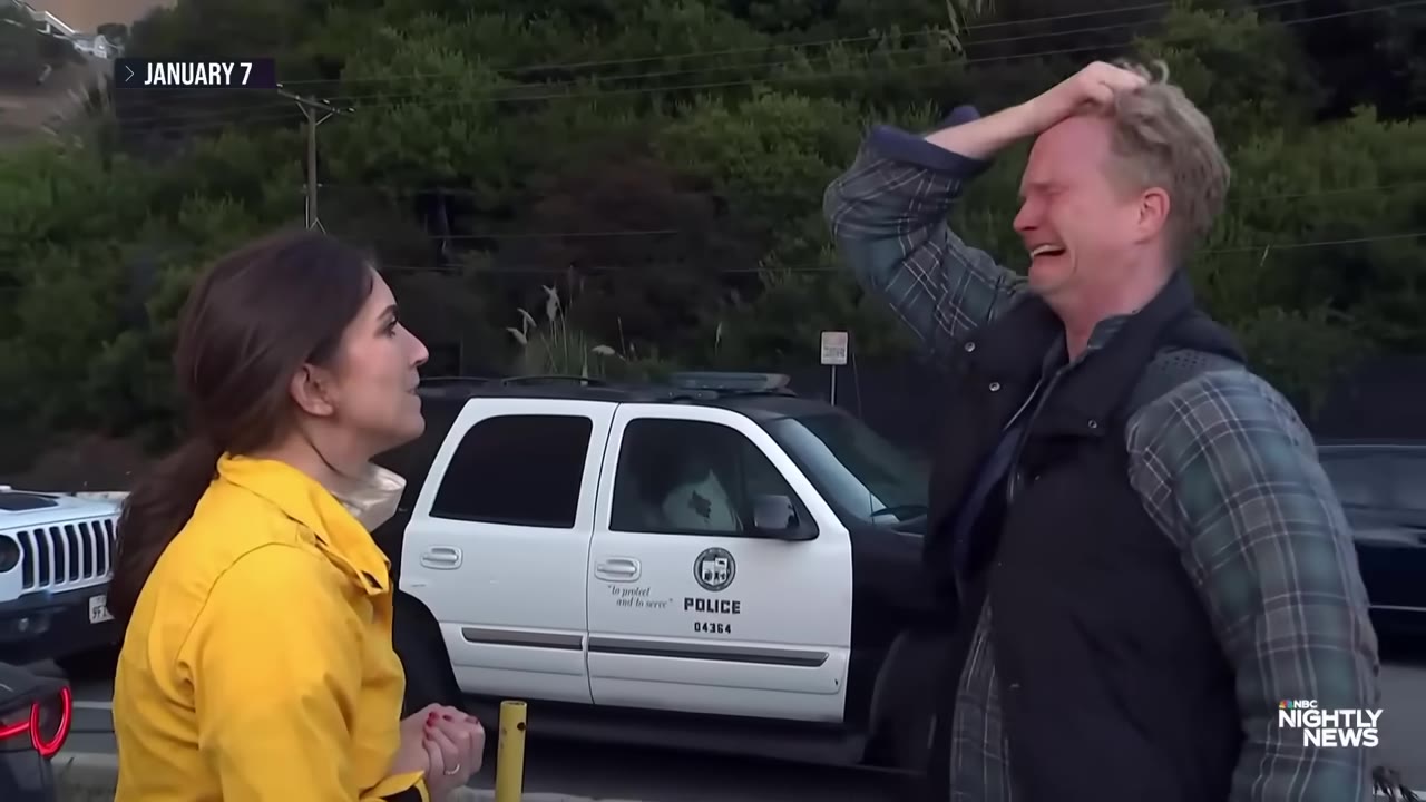 A man in a plaid shirt and vest stands outdoors with his hand on his head, crying with emotion, while a woman in a yellow jacket looks on next to a police vehicle.