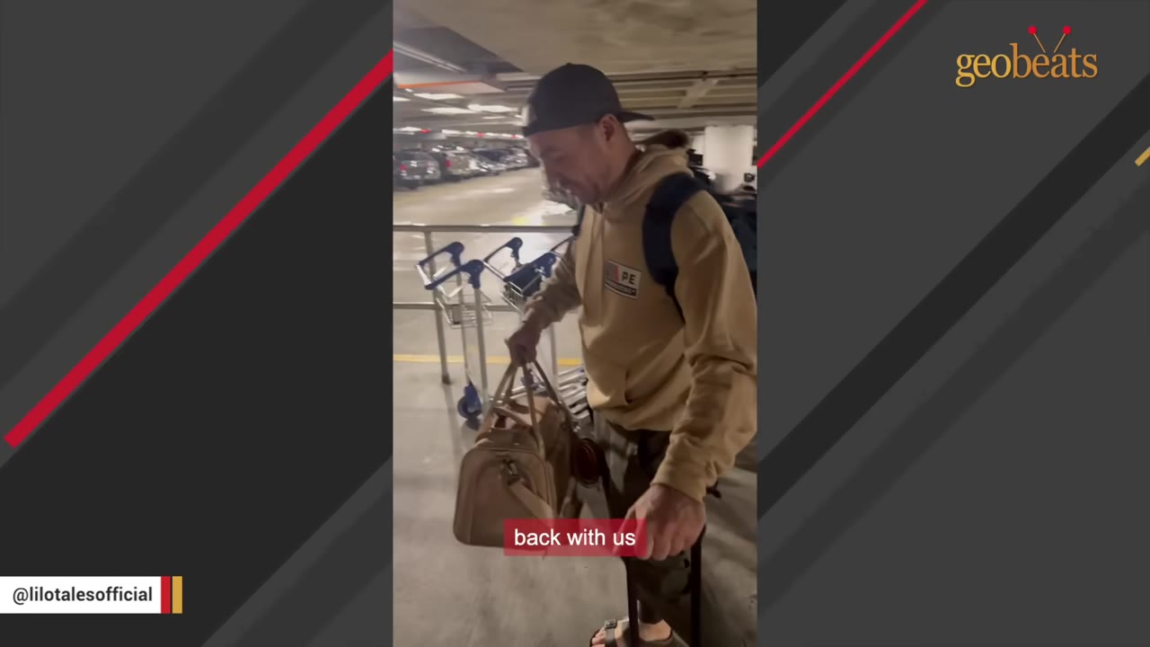 A man wearing a tan hoodie, a black baseball cap, and a backpack carries a tan pet carrier bag and a suitcase in a parking garage.