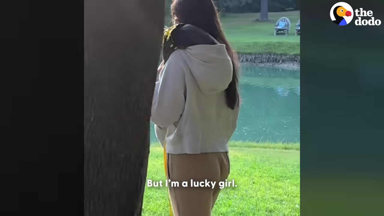 A young woman with long dark hair, wearing a light-colored hoodie and tan pants, stands near a pond with a dark cat wearing a yellow harness resting on her shoulder.